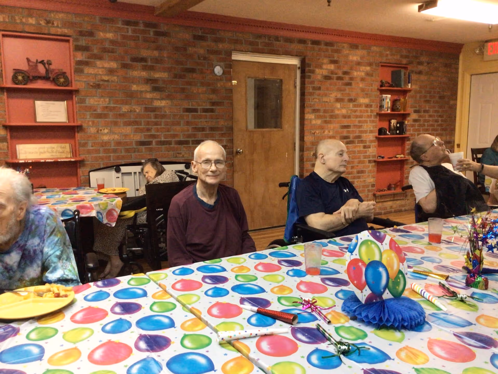 Several elderly residents sitting around a brightly decorated table with party decorations in a brick-walled communal dining room.