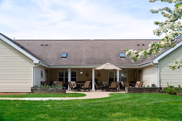 Outdoor patio area of a senior living facility with multiple tables and chairs arranged under a covered porch. There is a large umbrella over one table, green grass in the foreground, and light-colored siding on the building with a sloped roof and skylights.