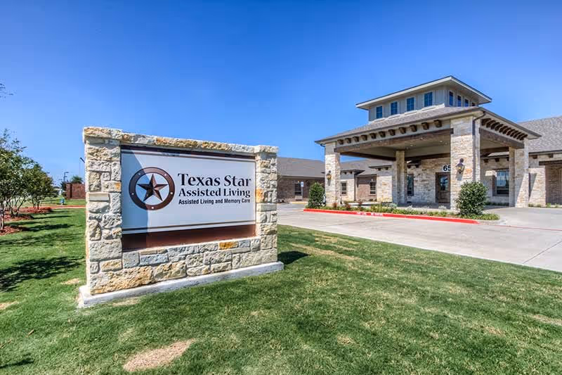 Exterior view of Texas Star Assisted Living facility showing a stone sign with the facility name and a building entrance with stone pillars and a covered driveway under a clear blue sky.