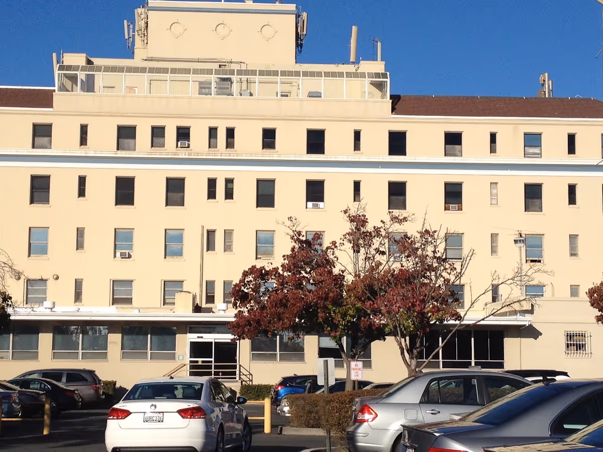 Exterior view of a multi-story beige building with several windows, a few trees with autumn-colored leaves in front, and a parking lot with multiple cars parked under a clear blue sky.