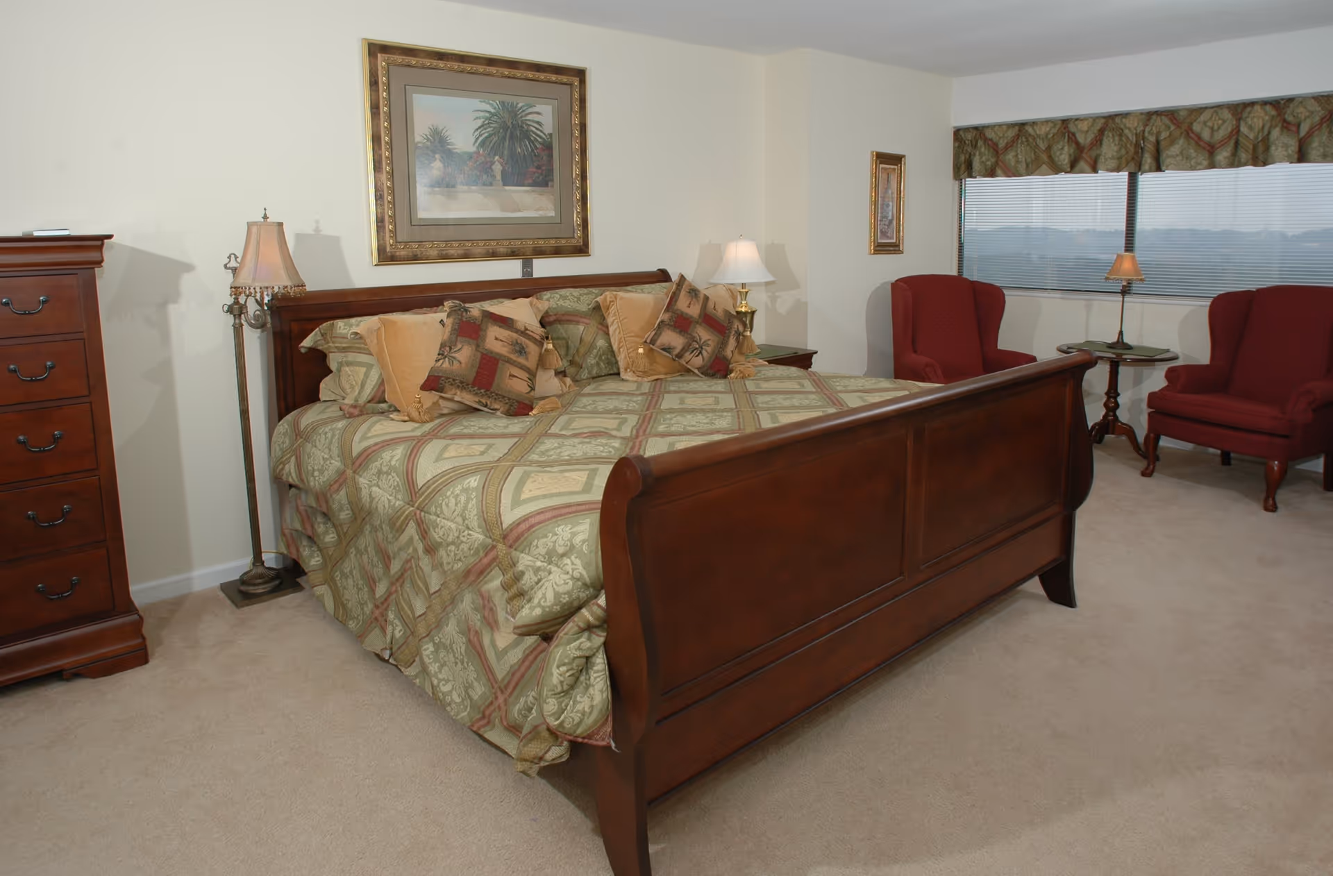 A bedroom with a large wooden bed covered with a patterned green and beige bedspread and multiple decorative pillows. There are two red upholstered armchairs with a small round table and lamp between them near a window with blinds and a valance. A wooden chest of drawers and two lamps are also visible, along with framed artwork on the walls.