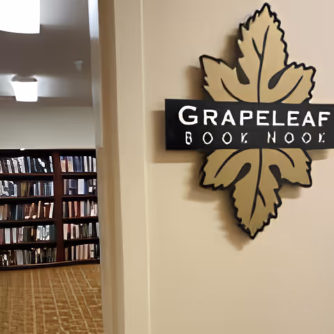 Interior view of a book nook area named Grapeleaf, featuring a decorative sign with a large leaf design mounted on a beige wall. In the background, there are bookshelves filled with books and a carpeted floor.