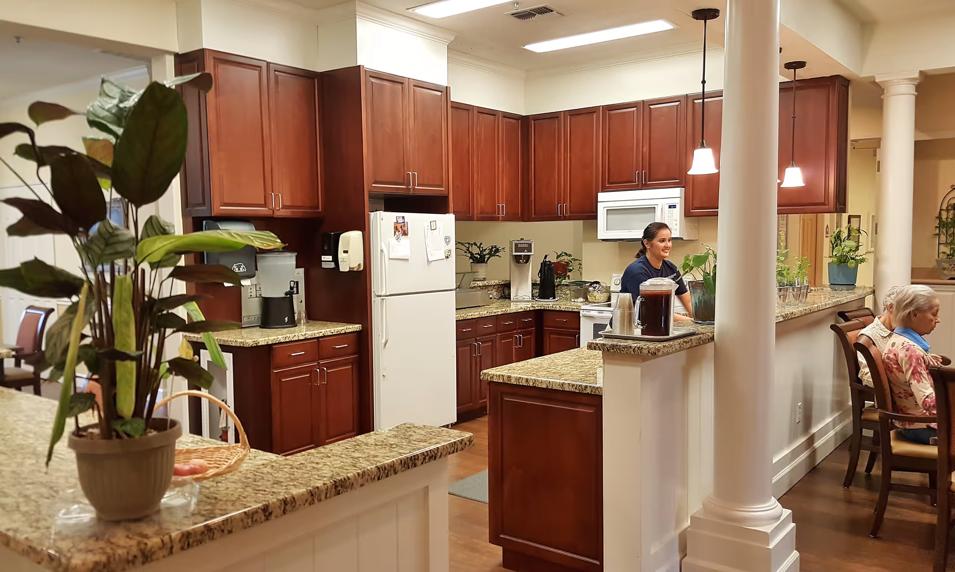 Communal kitchen and serving counter with cherry cabinets, granite countertops, a refrigerator, potted plants, a staff member behind the counter, and residents seated at a nearby table.