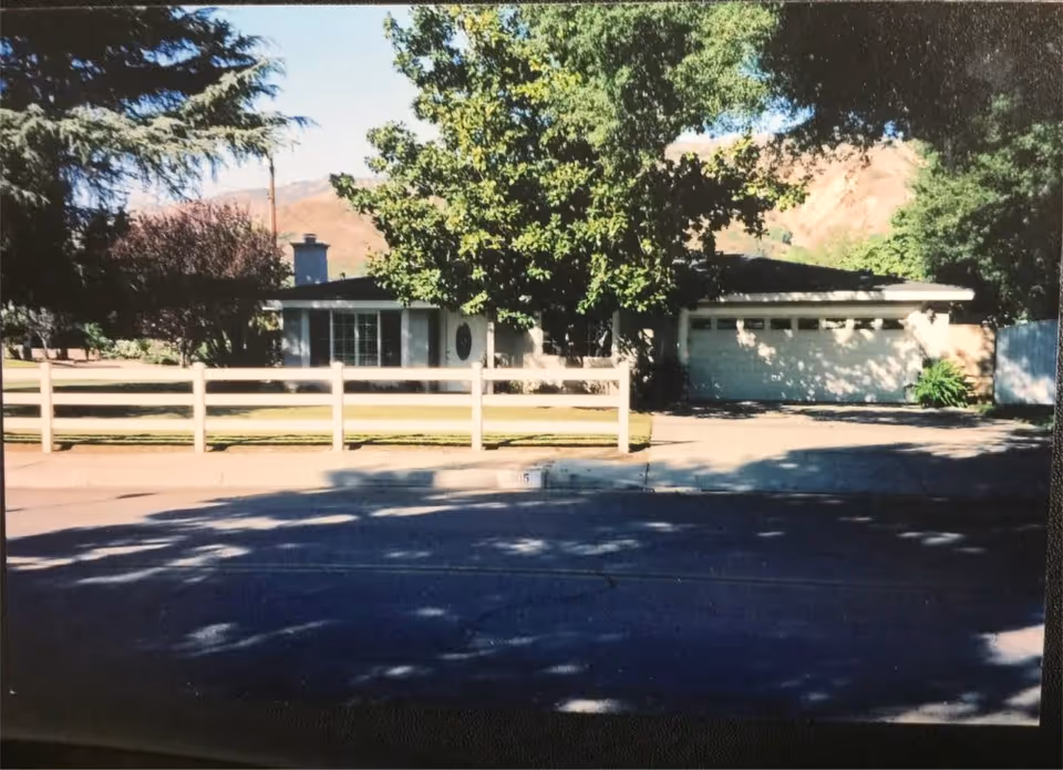 Front exterior view of a single-story house with a white fence, a driveway leading to a two-car garage, and several trees providing shade. Mountains are visible in the background under a clear sky.