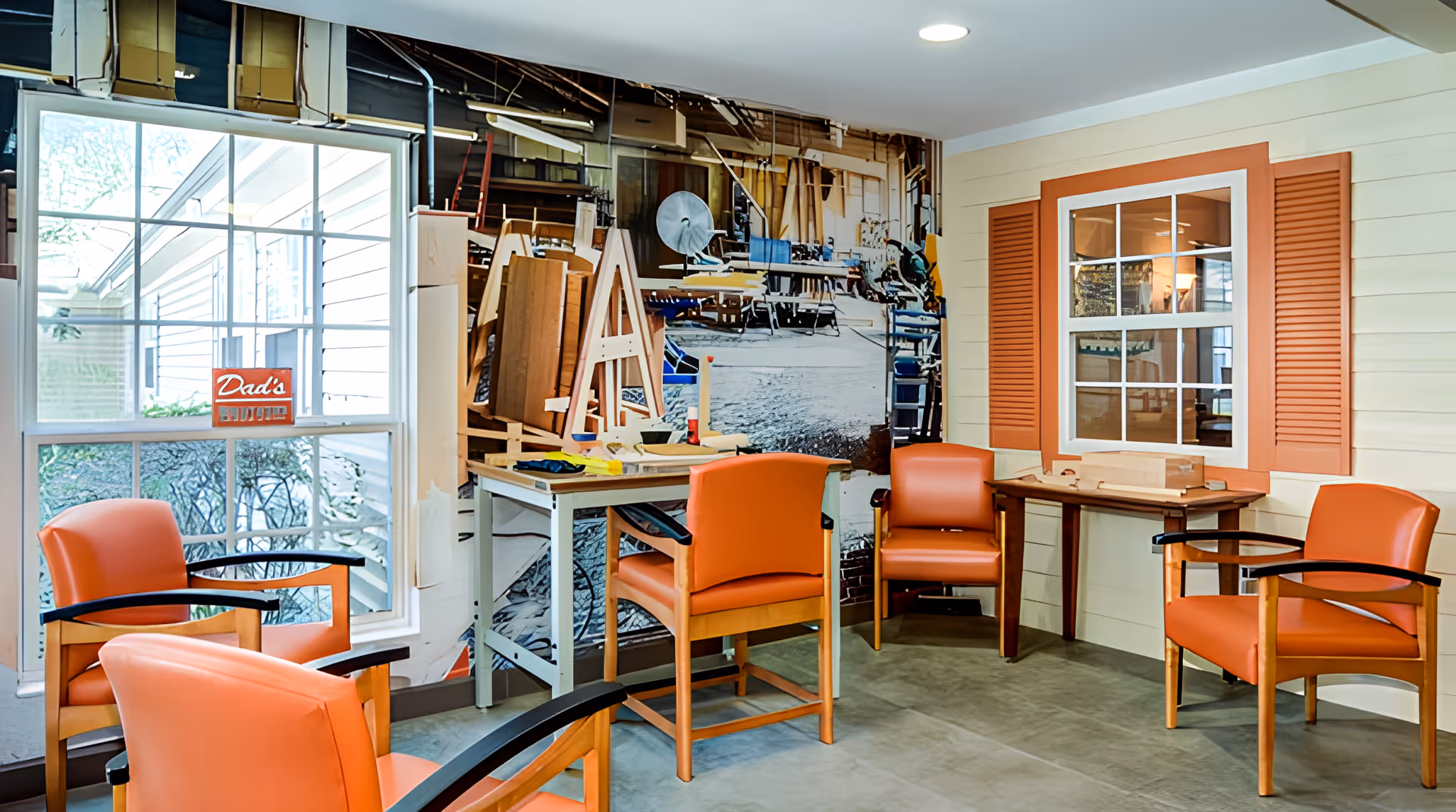 A cozy seating area with five orange chairs arranged around two small tables. The room features a large window with a sign that says 'Dad's' and a wall mural depicting a workshop or garage scene. The walls are light-colored with a decorative window frame and orange shutters.