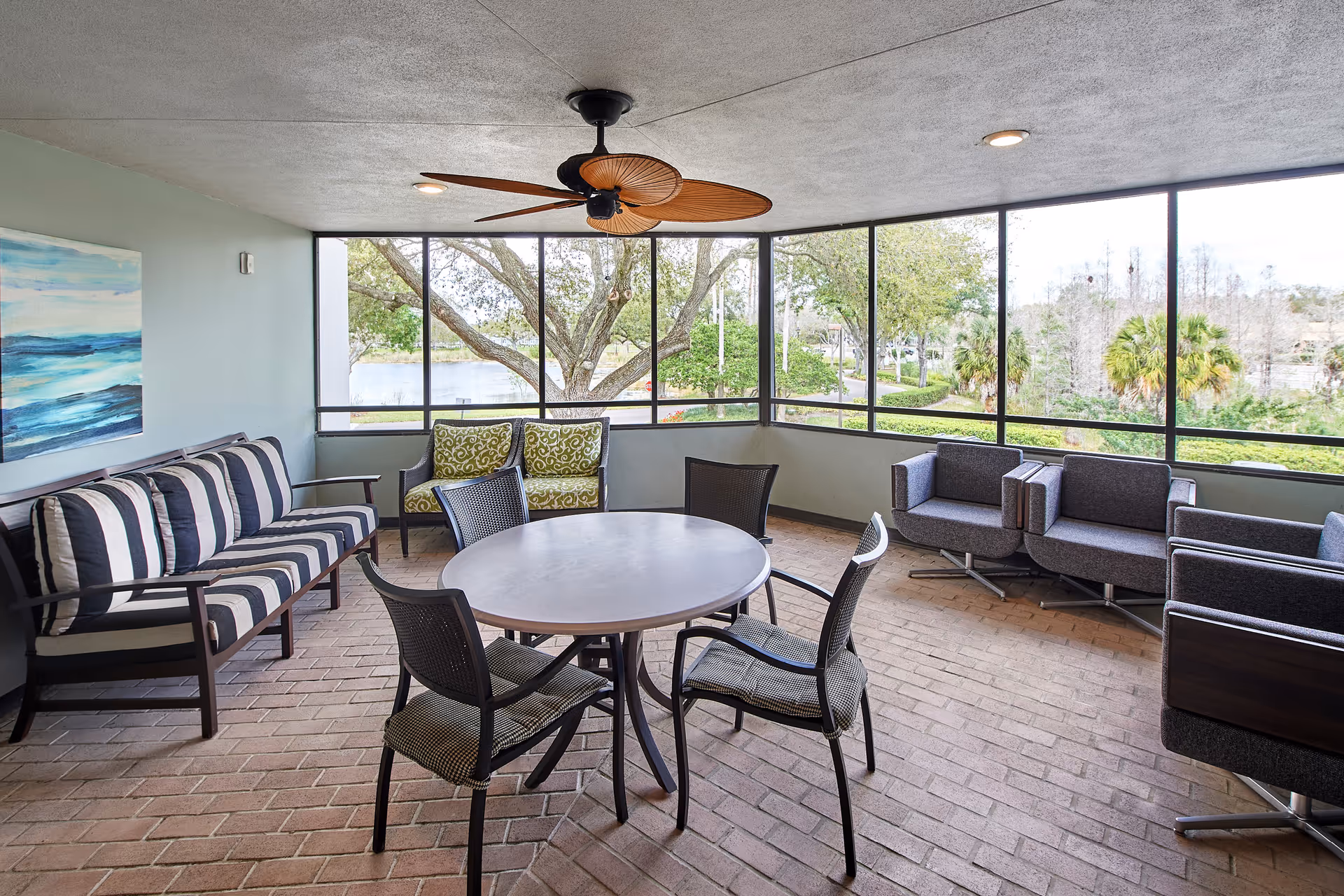 A bright enclosed patio area with large windows overlooking trees and greenery. The space features a round table with four chairs in the center, a striped cushioned bench along one wall, two green patterned armchairs, and three modern gray armchairs arranged on the right side. A ceiling fan with large wooden blades hangs from the ceiling, and a blue and white abstract painting is mounted on the left wall.