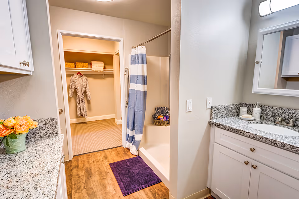 A bright bathroom with white cabinets and granite countertops. There is a sink with soap and lotion dispensers, a mirror above it, and a shower with a blue and white striped curtain. A purple bath mat is on the wooden floor in front of the shower. Through an open door, a walk-in closet is visible with a hanging robe and shelves holding baskets and towels.