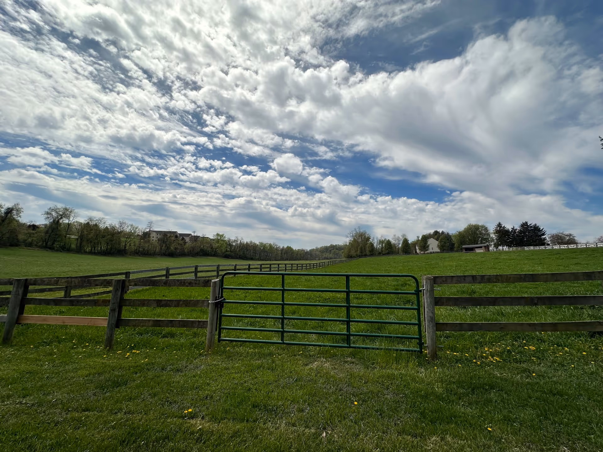 A green pasture enclosed by wooden fences with a metal gate in the center. The sky above is partly cloudy with patches of blue. Trees and a few buildings are visible in the distance.
