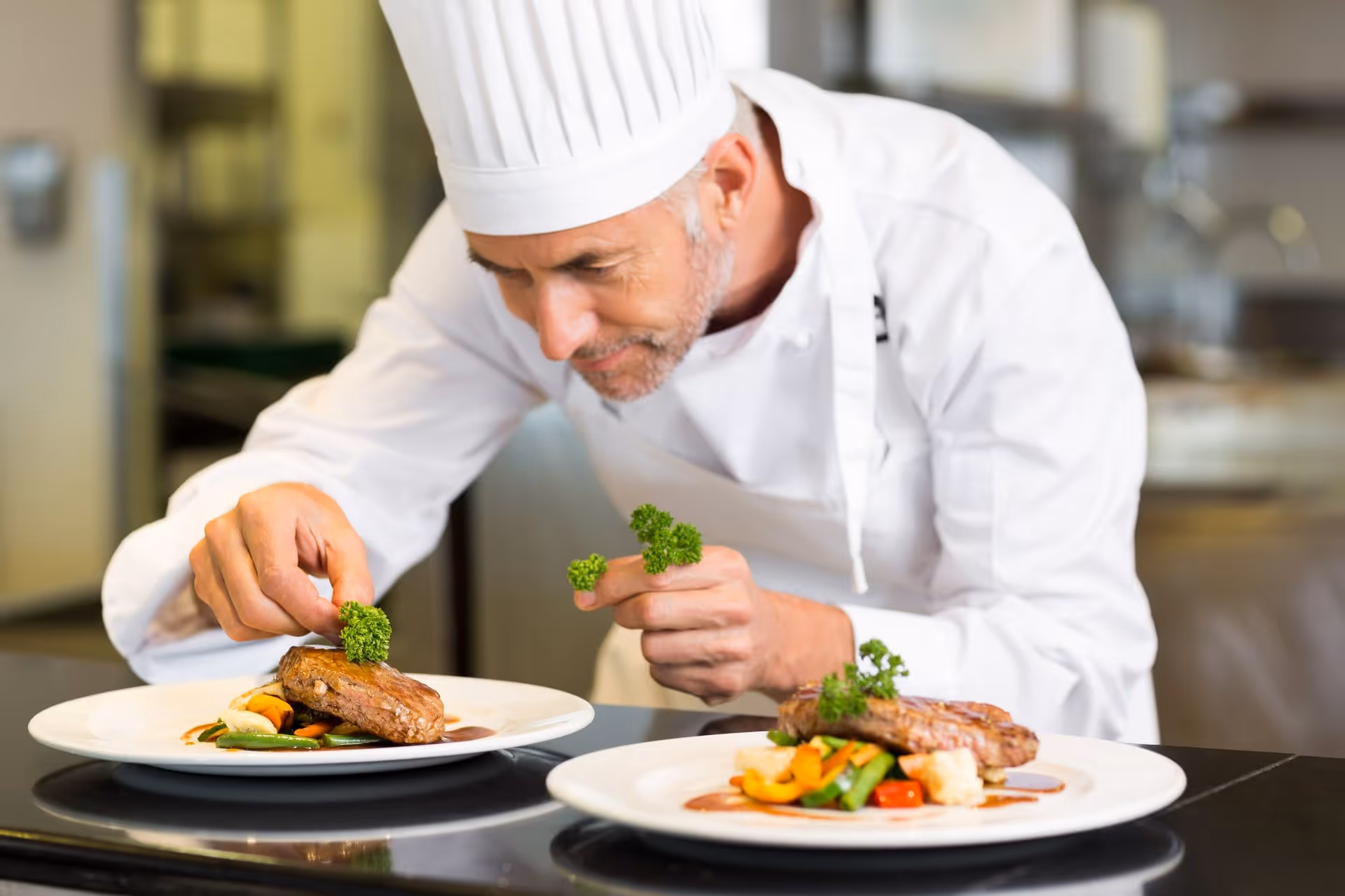 A chef wearing a white uniform and hat carefully garnishes two plates of food with fresh green herbs in a professional kitchen setting.