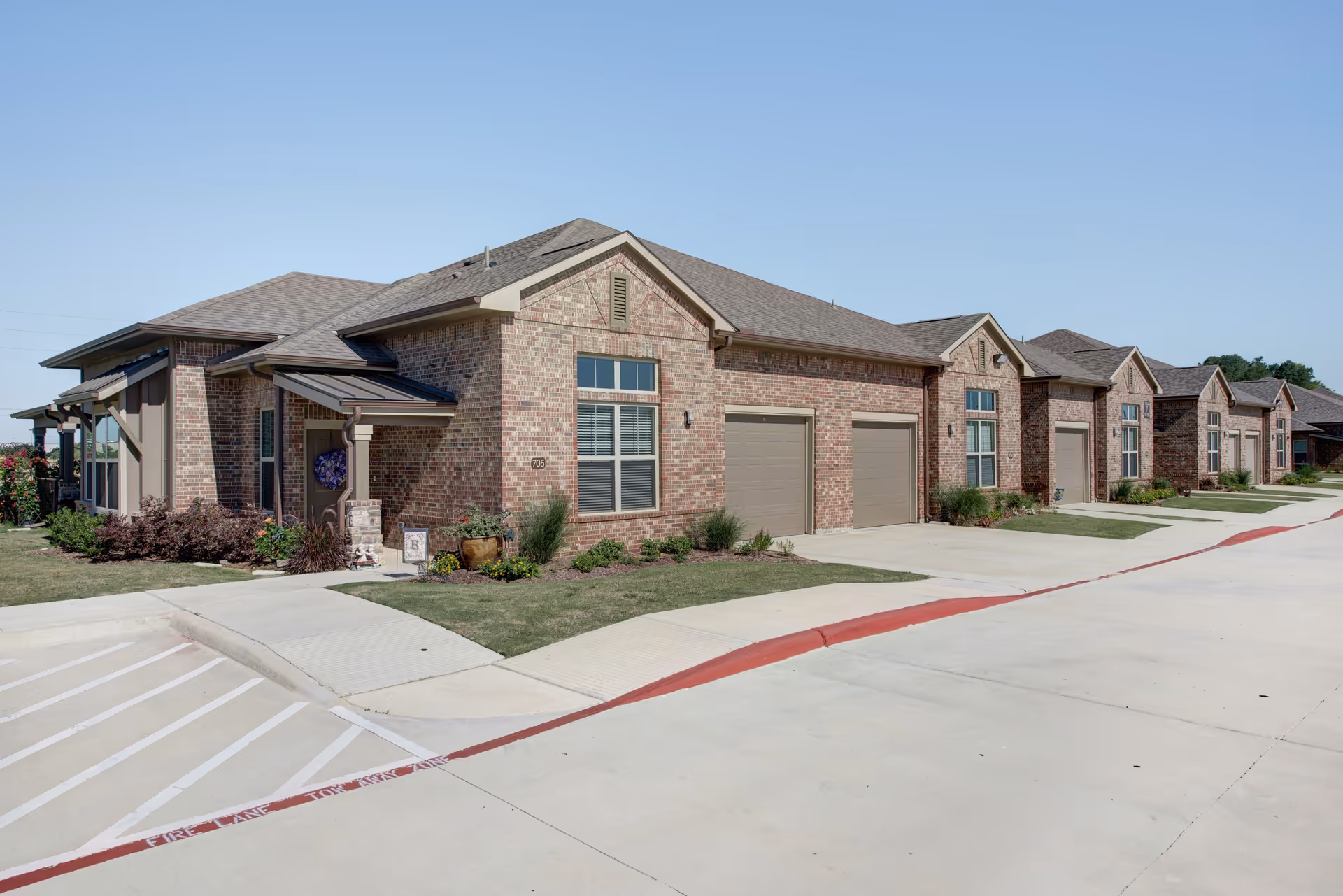Exterior view of a single-story brick building with multiple garage doors and windows, surrounded by a concrete driveway and landscaped with grass and shrubs under a clear blue sky.