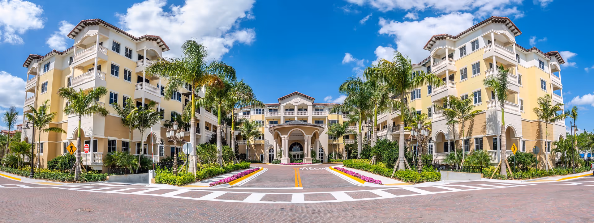 Front exterior view of The Palace at Weston, a large multi-story building with yellow and white walls, red-tiled roof, balconies, palm trees, and a covered entrance under a blue sky with scattered clouds.