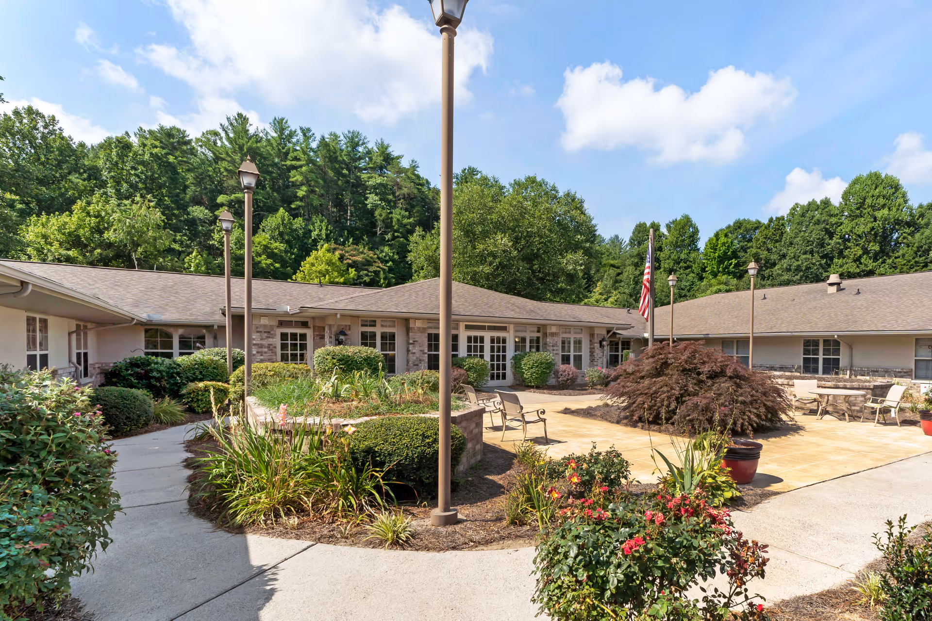 Outdoor courtyard area at Brookdale Lenoir featuring a paved walkway, benches, landscaped bushes and plants, lamp posts, and an American flag, surrounded by single-story building structures and trees in the background under a partly cloudy sky.