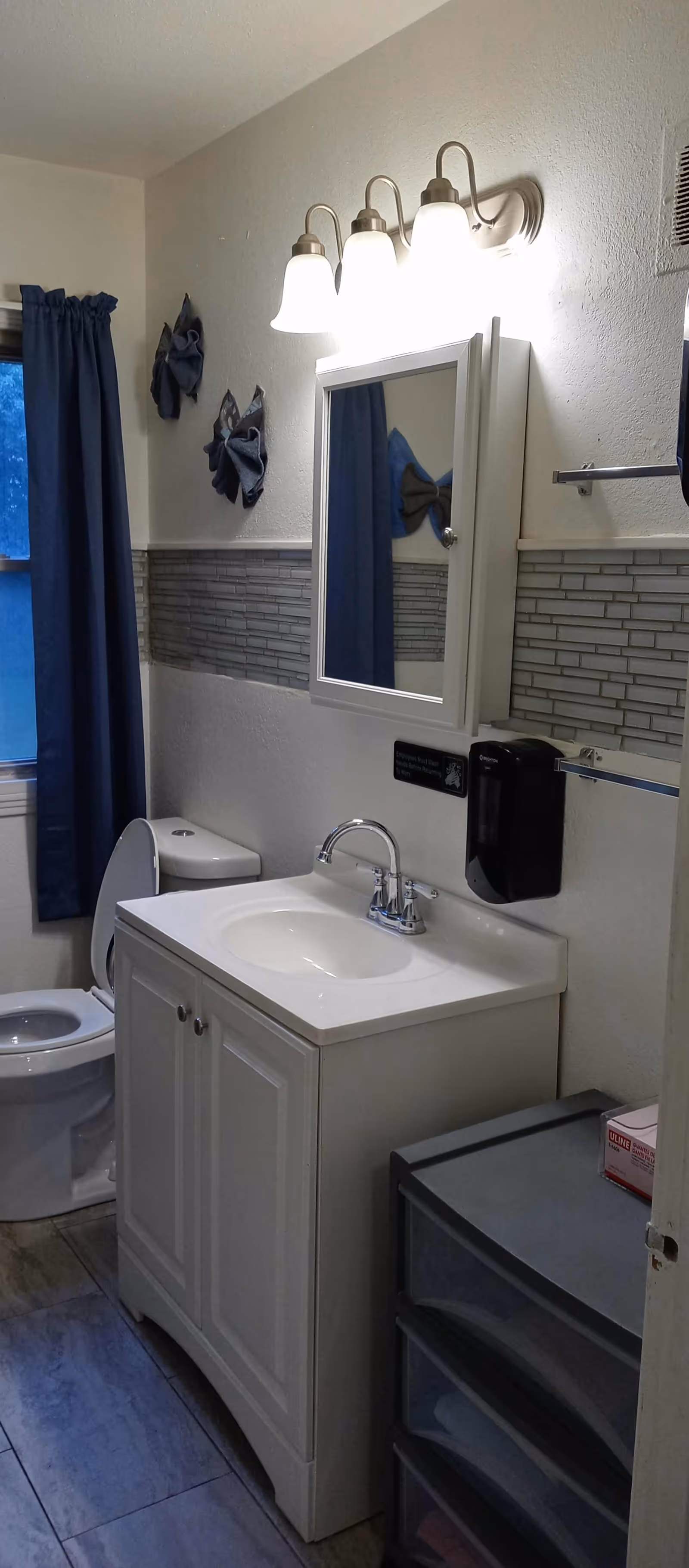 A bathroom with a white vanity cabinet and sink, a wall-mounted mirror with three light fixtures above it, a toilet with the lid open, a window with dark blue curtains, and a small plastic drawer unit. The walls have a decorative tile strip and two towel holders.