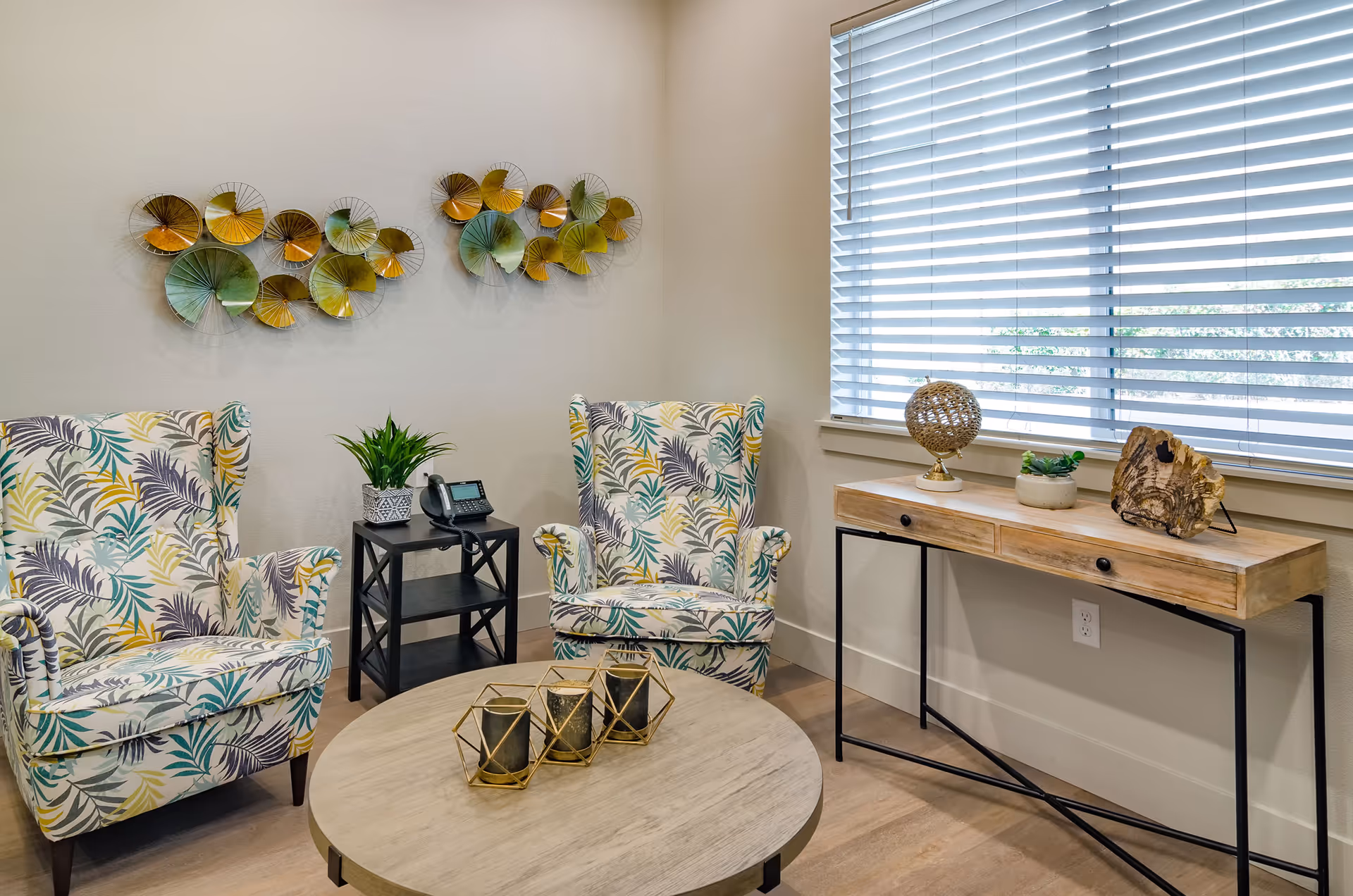 A bright sitting area with two patterned armchairs, a round coffee table, decorative wall art, and a console table beneath a window.