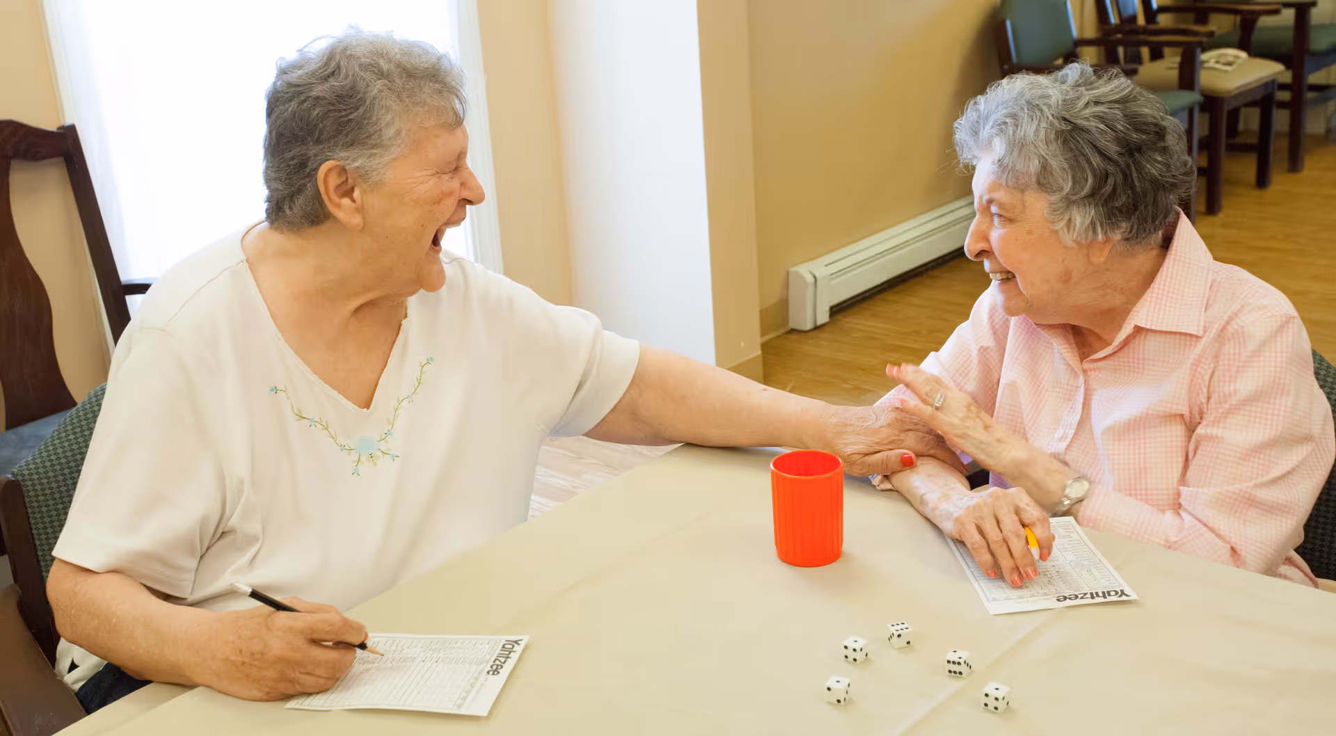 Two elderly women sitting at a table playing a game with dice and scorecards, smiling and holding hands in a warm, friendly interaction inside a room with wooden flooring and chairs.