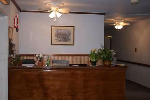 Reception desk in a dim lobby with plants, framed art, and ceiling fans.