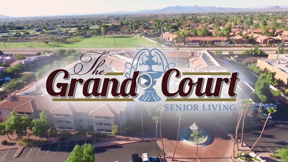 Aerial view of a senior living facility named Grand Court, showing multiple buildings with red-tiled roofs, surrounded by trees and parking areas, with a fountain in the circular driveway at the entrance. The background includes a golf course and distant mountains under a clear sky.