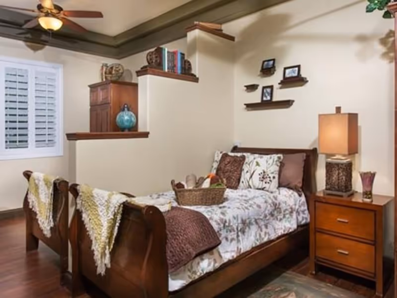 A cozy bedroom with a wooden bed featuring floral bedding and decorative pillows. A basket with bread and fruit is placed on the bed. Next to the bed is a wooden nightstand with a lamp and a small vase. The room has wooden flooring, a ceiling fan with a light, and a window with white shutters. There are small shelves on the wall above the bed displaying framed photos.