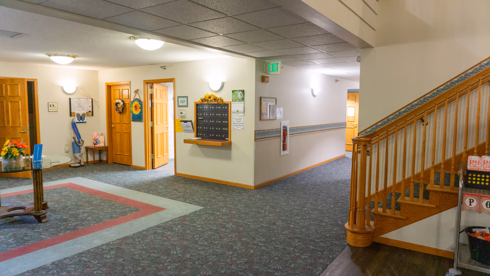 Interior view of a senior living facility hallway with carpeted floors and wooden doors. There is a wooden staircase on the right side, a glass table with a flower arrangement on the left, and a wall-mounted mailbox unit in the center. The walls are decorated with framed pictures and a sunflower wreath, and there is an exit sign above a door in the background.