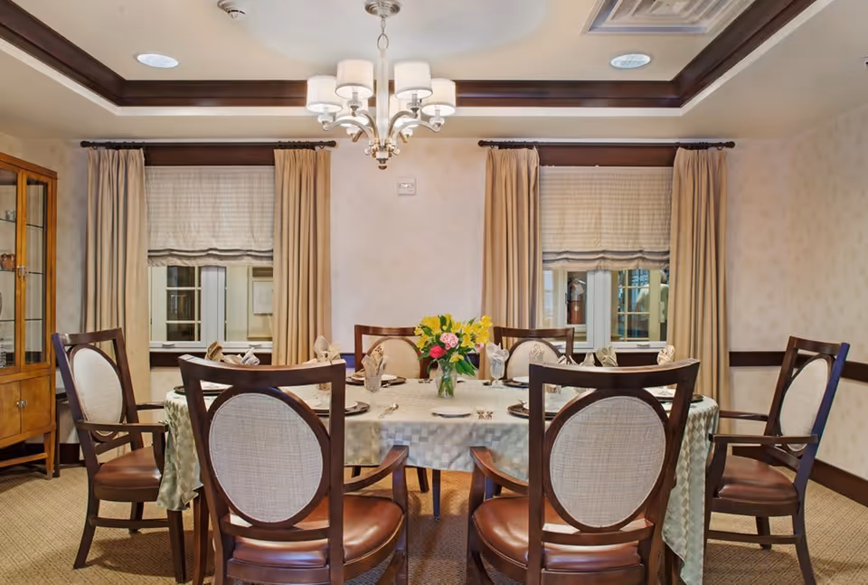 A formal dining room with a round table covered by a checkered tablecloth, set with plates, napkins, and silverware. Six wooden chairs with cushioned seats and backs surround the table. A vase with colorful flowers is placed in the center. The room has two windows with beige curtains and Roman shades, a wooden cabinet with glass doors on the left, and a chandelier hanging from a tray ceiling.