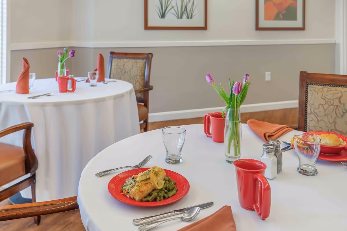 Round dining tables set with plates of food, red mugs, glassware, folded napkins, and small vases of tulips in a bright dining room.