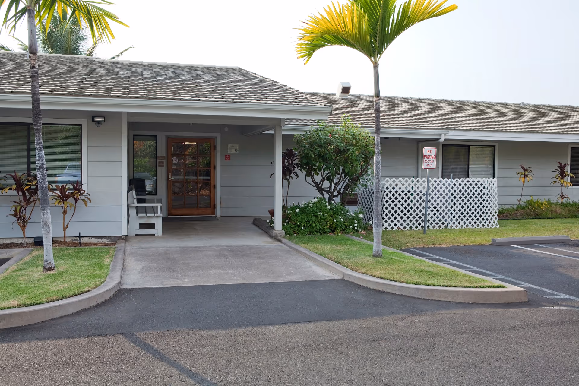Exterior view of a single-story building with a covered entrance, a wooden door with glass panels, palm trees, and a small garden area. There is a parking area with a sign that reads 'No Parking Doctor Only'.