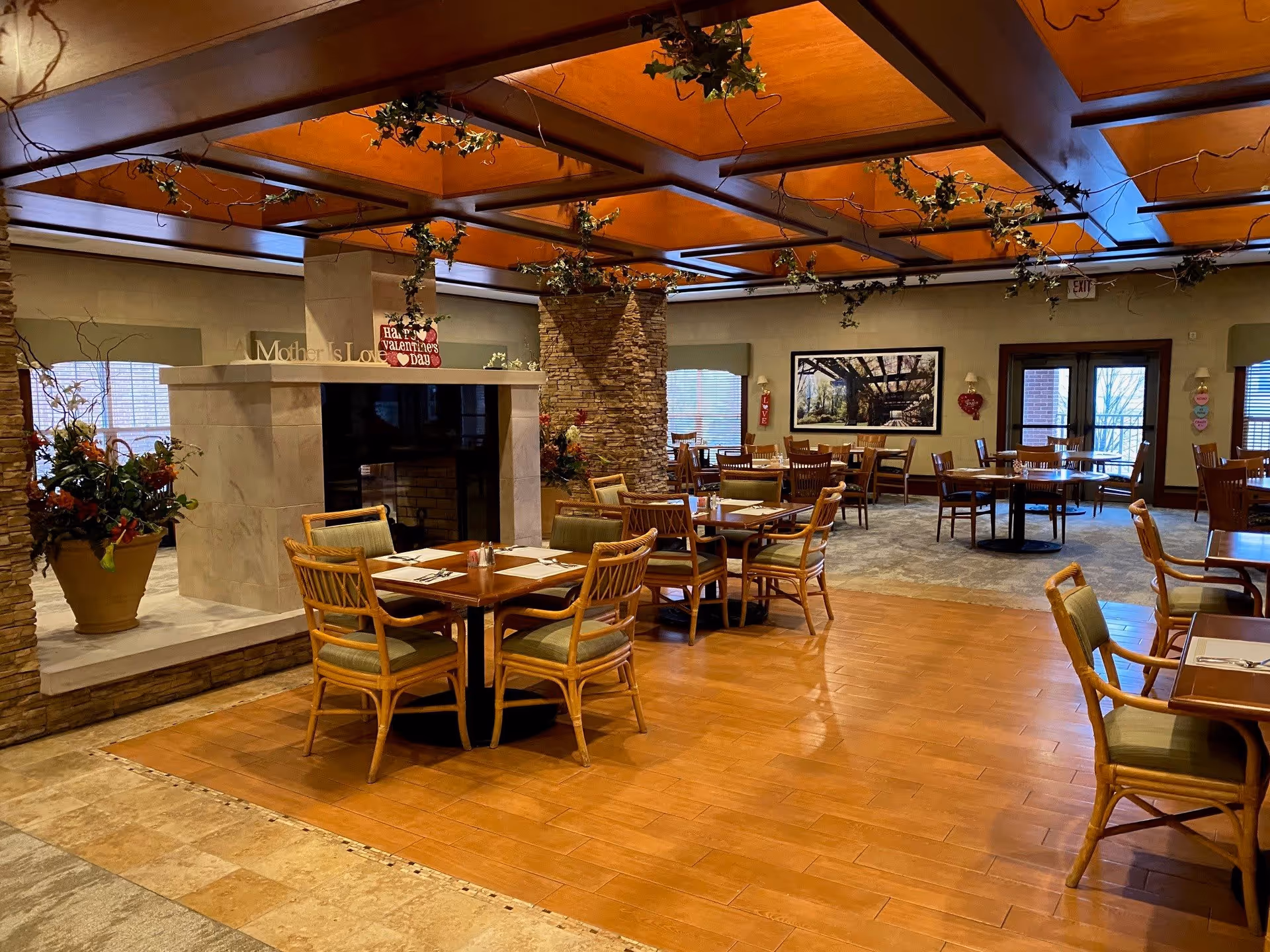 A spacious dining room with wooden tables and chairs arranged neatly. The room features a stone fireplace in the center with a 'Happy Valentine's Day' sign on it. The ceiling has wooden beams decorated with green vines. Large windows allow natural light to fill the room, and there are various floral arrangements and wall decorations.