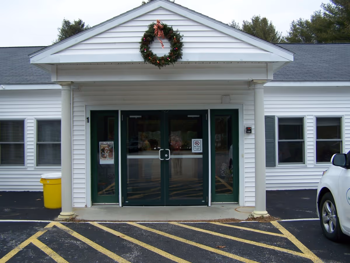 Entrance of The Lodges Care Center with white siding, green double doors, a Christmas wreath above the doors, and a yellow trash bin to the left. There is a white car parked on the right side and yellow striped markings on the asphalt in front of the entrance.
