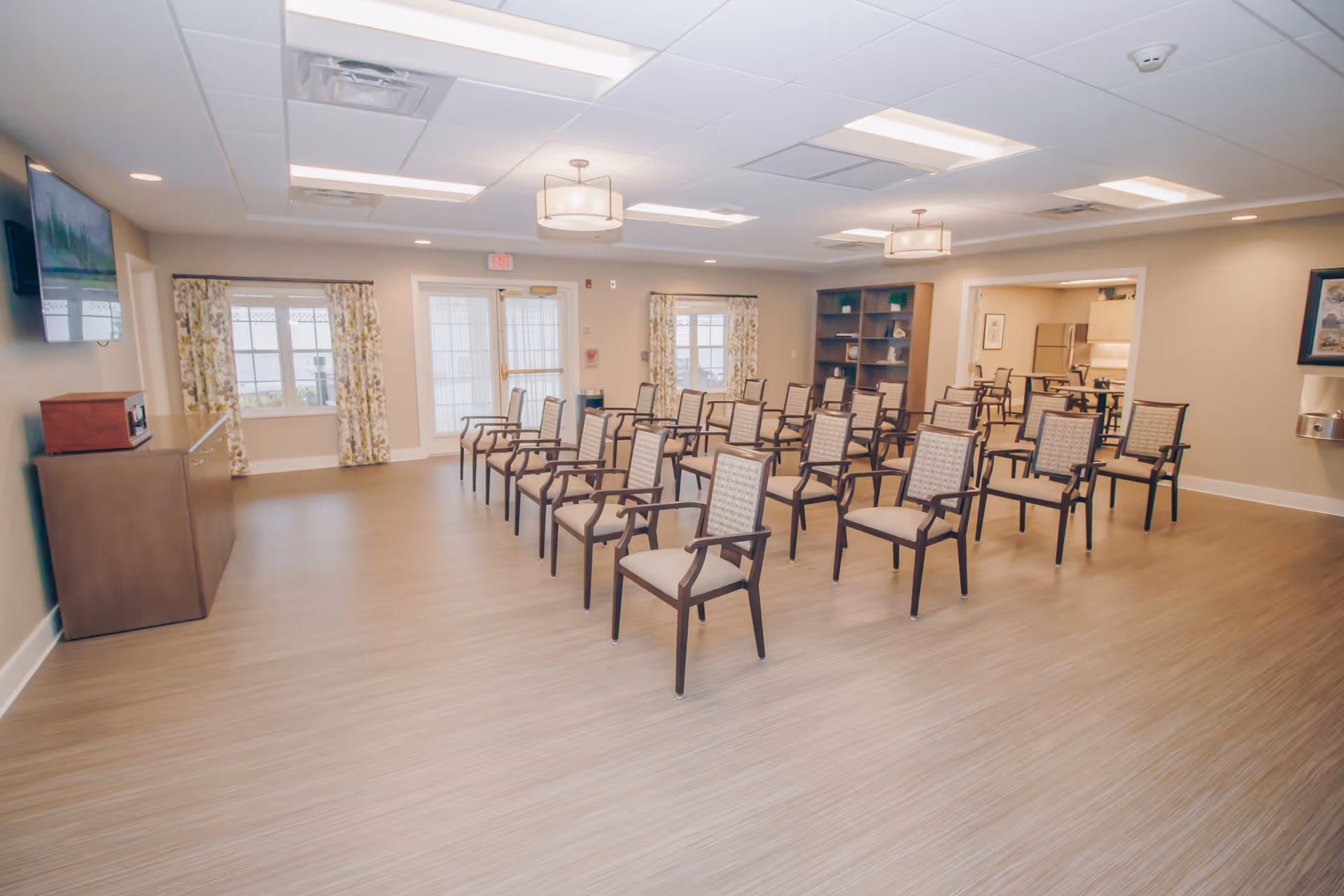 A spacious room with several rows of wooden chairs with cushioned seats and backs arranged facing a wall-mounted television. The room has light-colored walls, wood flooring, two large windows with floral curtains, and two ceiling light fixtures. There is a wooden cabinet on the left side and a bookshelf on the right side near an open doorway leading to another room with kitchen appliances.