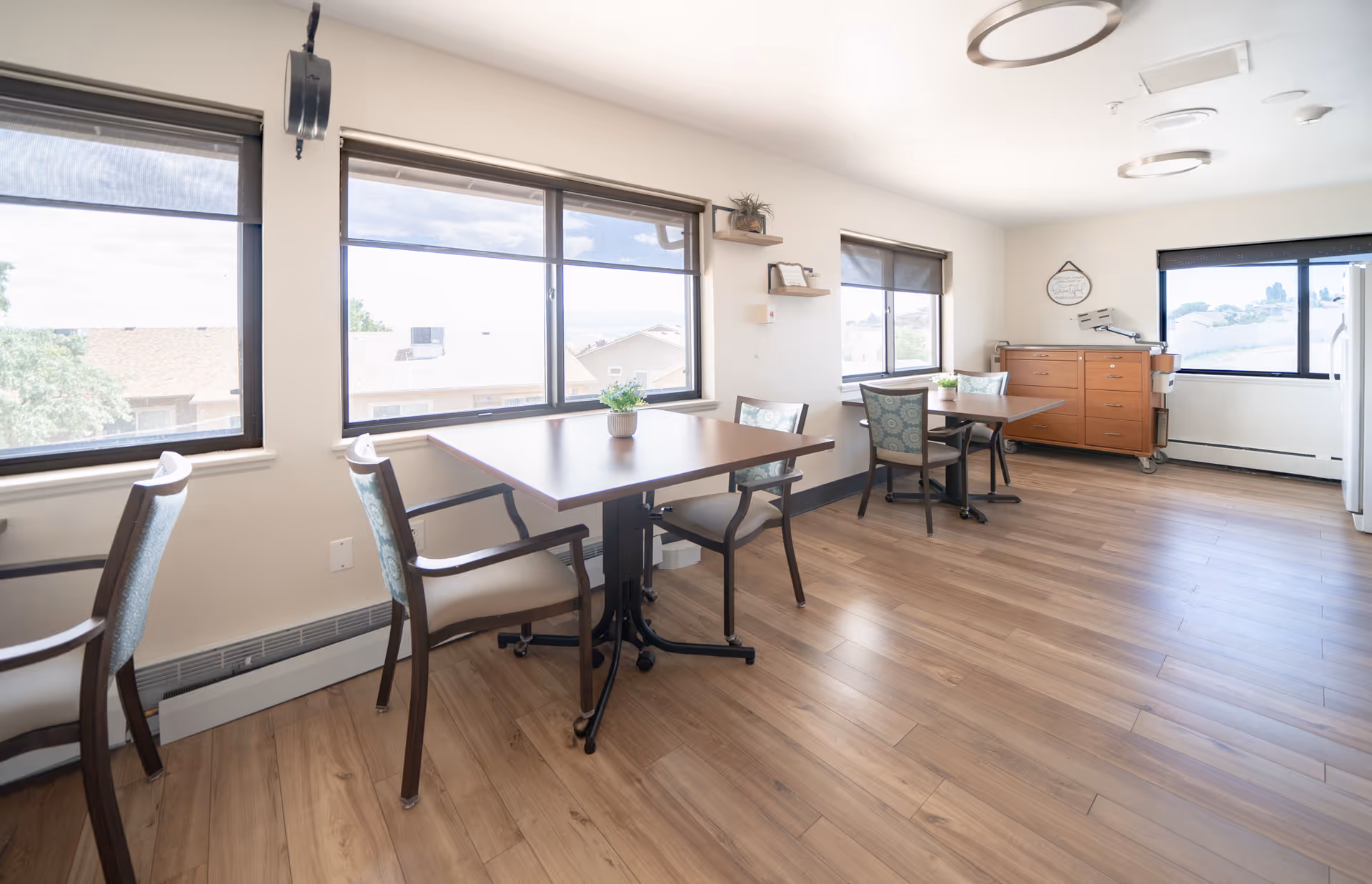 A bright dining room with wooden floors, several tables with chairs, large windows with roller shades letting in natural light, and a wooden sideboard against the far wall with decorative items on top.