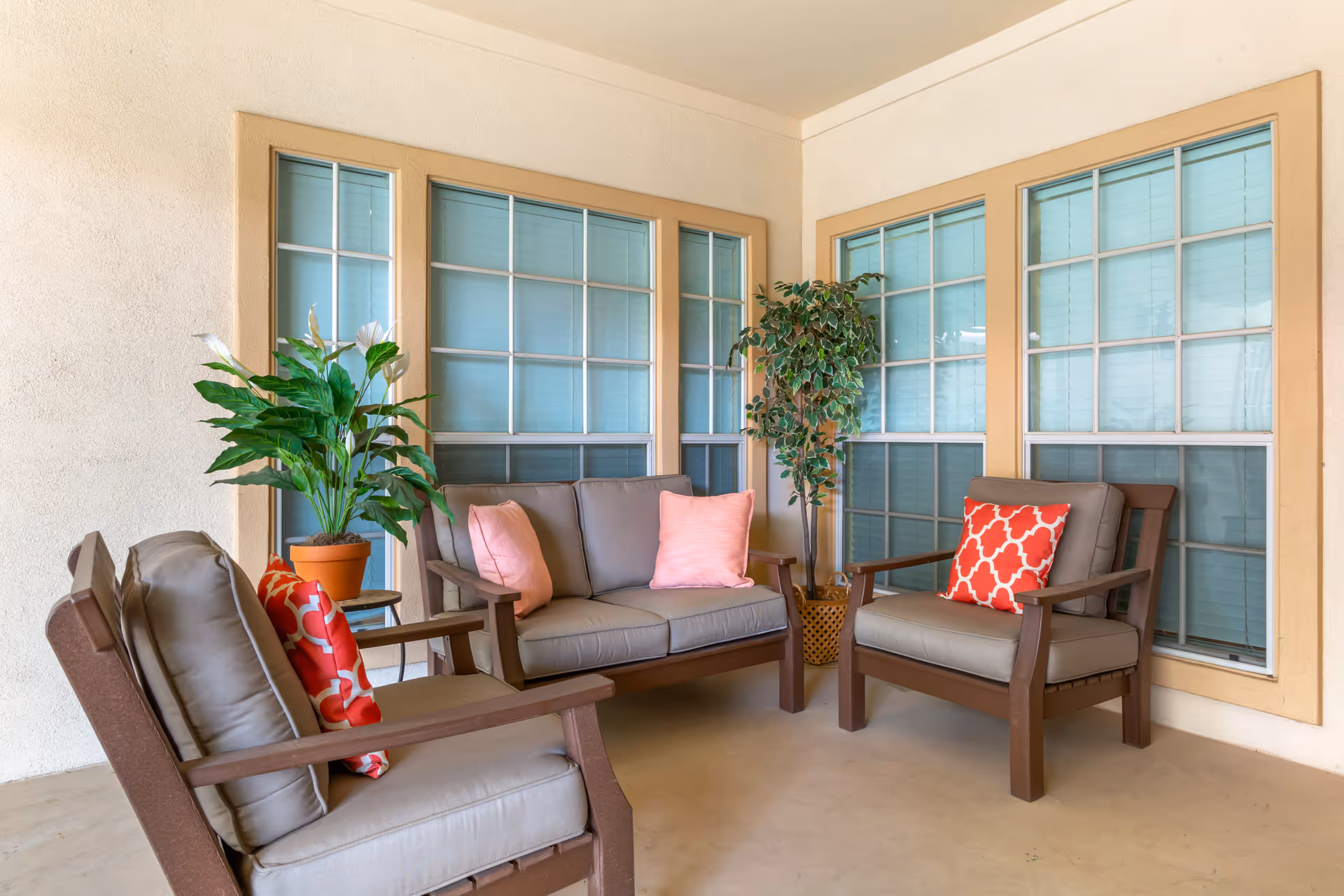 A cozy indoor seating area with three cushioned wooden chairs and a loveseat, each adorned with colorful throw pillows. The space is decorated with two potted plants and surrounded by windows with beige trim and blue blinds.