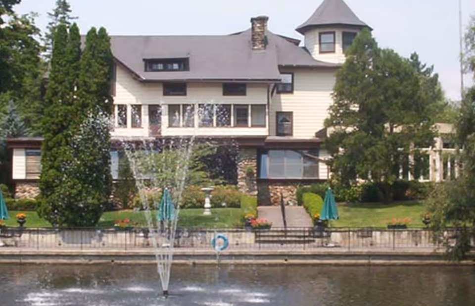A large, multi-story house with a turret and multiple windows, surrounded by tall trees and greenery. In front of the house is a pond with a water fountain spraying water upwards. There are stairs leading from the house to the pond area, with green umbrellas and flower pots on either side.