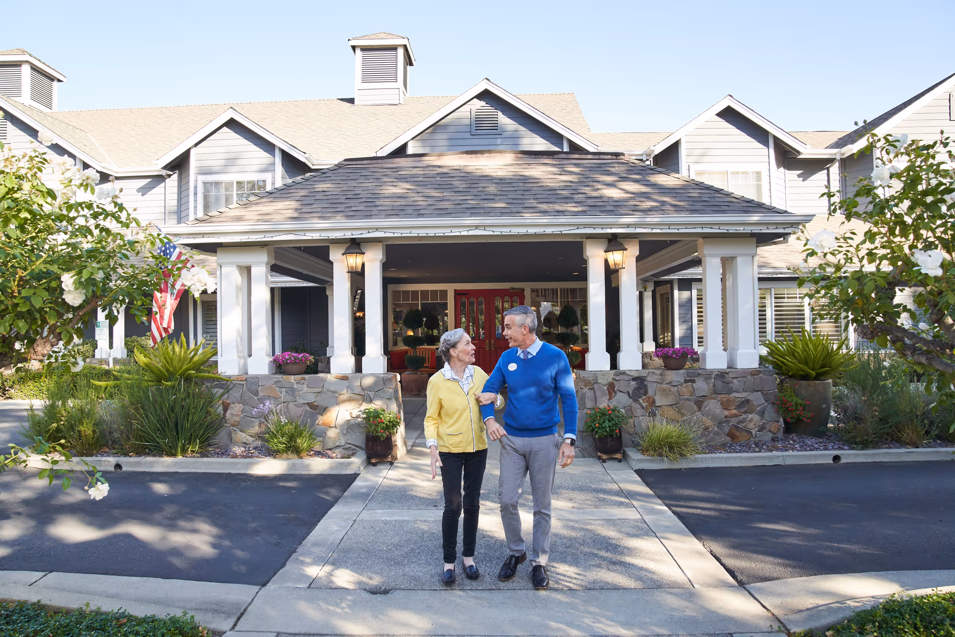 An elderly couple walking arm in arm on a sidewalk in front of a senior living facility entrance with a covered porch, stone pillars, and a well-maintained garden on either side.