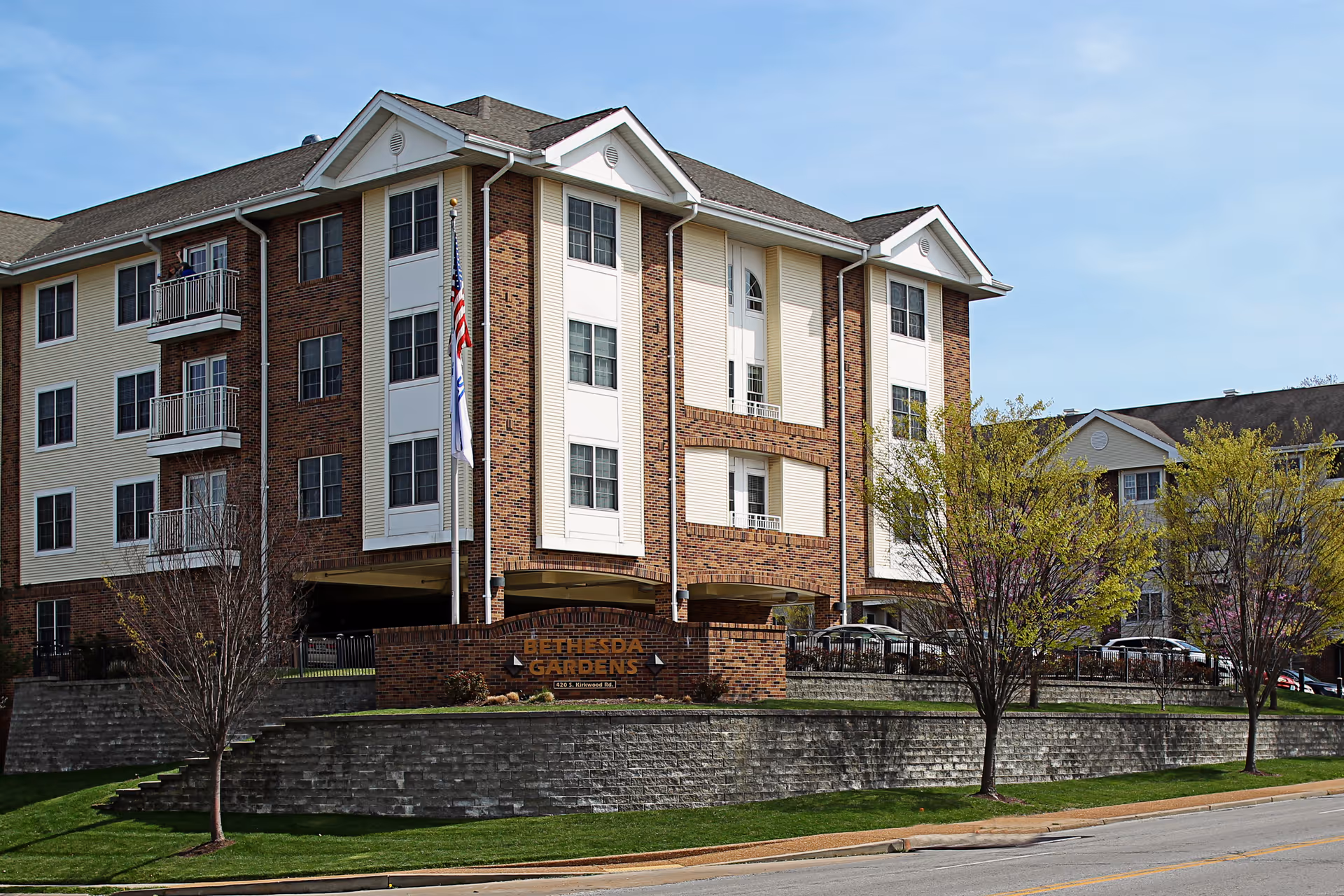 Exterior view of a multi-story brick and white panel building with balconies, surrounded by trees and a stone retaining wall. A sign in front reads 'Bethesda Gardens' with an address below. Two flagpoles with flags are positioned near the sign.