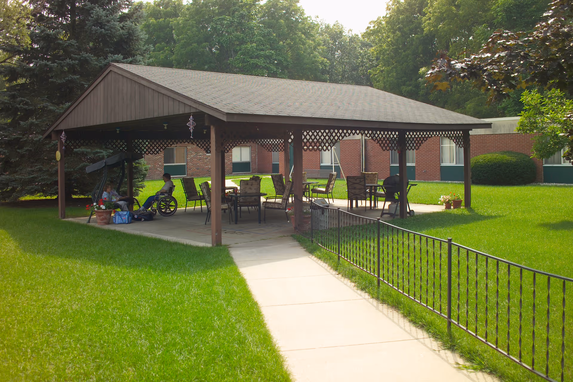 Covered outdoor pavilion with tables and chairs on a grassy courtyard, a person in a wheelchair under the shelter and a brick building in the background.
