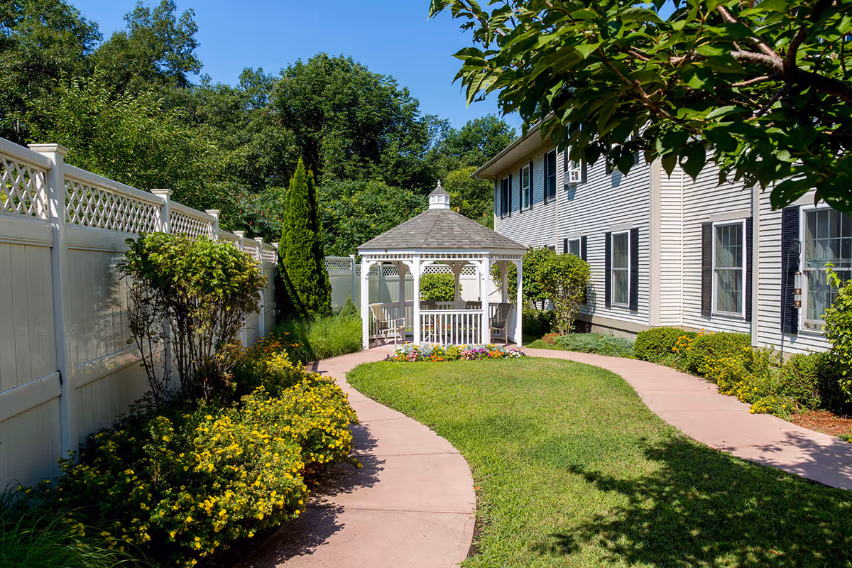 A sunny outdoor garden area with a curved paved walkway leading to a white gazebo with seating. The garden is bordered by a white fence on the left and a beige building with multiple windows on the right. There are green bushes, yellow flowers, and trees surrounding the area under a clear blue sky.