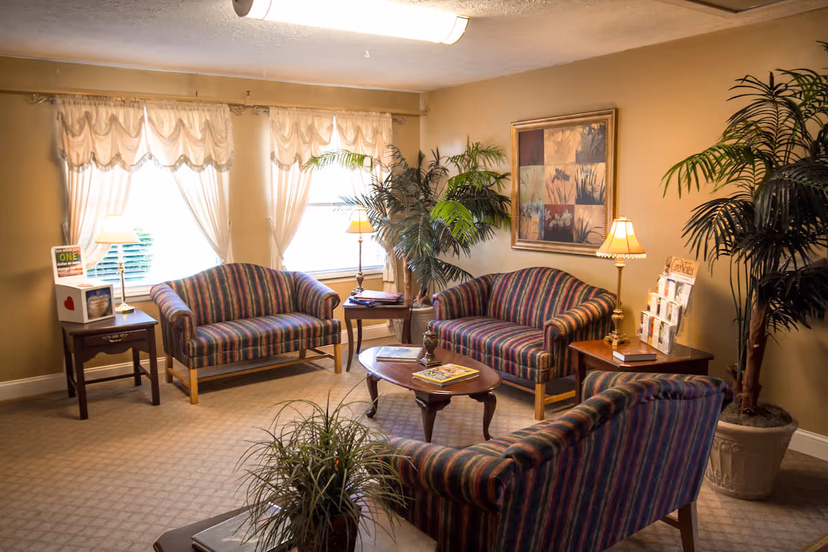 A cozy living room area with three striped sofas arranged around a wooden coffee table. The room has beige walls and carpet, two large windows with sheer curtains, two table lamps on side tables, potted plants, and a framed artwork on the wall.