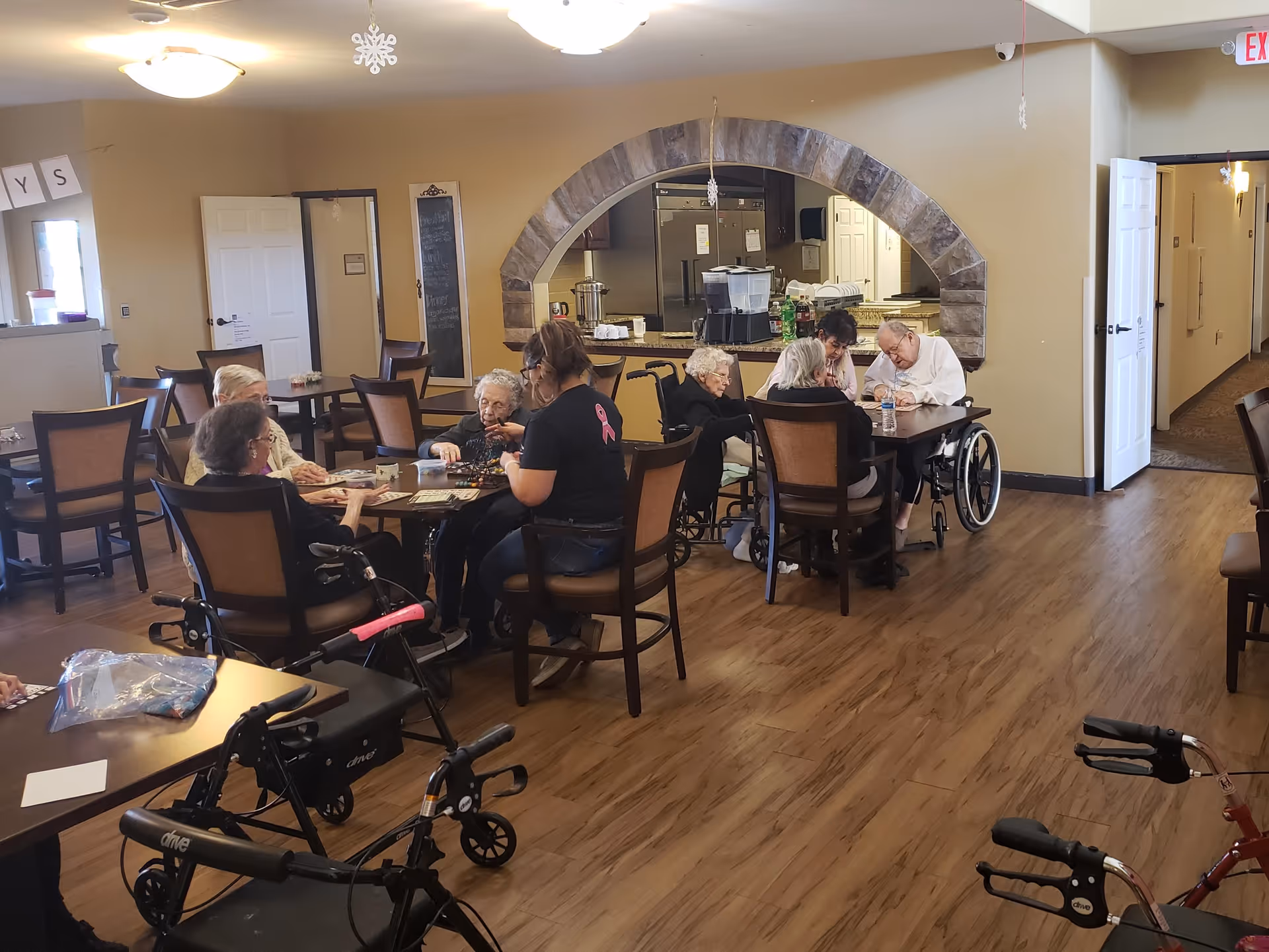 A group of elderly people sitting around tables in a common area of an assisted living facility, engaging in activities with the help of a caregiver. The room has wooden floors, beige walls, and an arched opening leading to a kitchen area in the background. Several walkers are visible in the foreground.