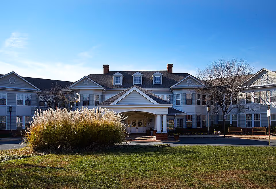 Front exterior view of a large senior living facility building with white siding, multiple windows, and a covered entrance with columns. There is a grassy area and ornamental grasses in front of the building under a clear blue sky.