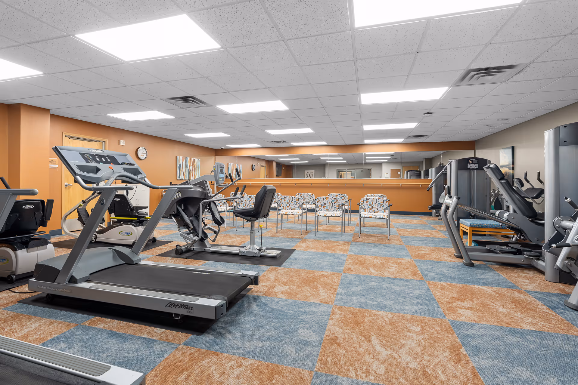 Carpeted fitness room with treadmills, exercise machines, and a row of chairs in front of a mirrored wall.