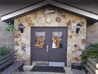 Stone facade entrance of a building with double dark brown doors decorated with autumn-themed wreaths. Above the doors is a small sign that says 'Welcome'. Two black wall-mounted lantern lights flank the doors. There are benches on either side of the entrance and some greenery, including a hanging plant on the left and bushes on the right.