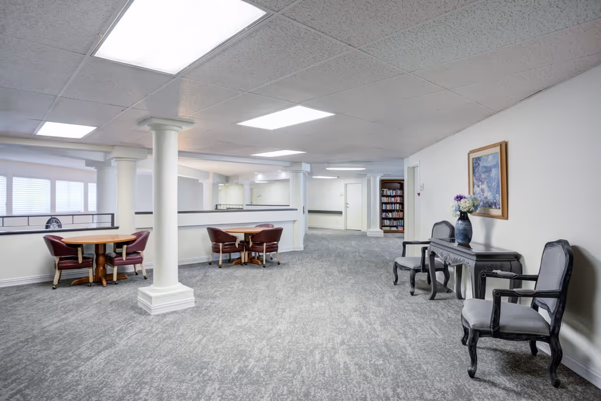 A spacious, well-lit common area with gray carpet and white walls. The room features two round wooden tables each surrounded by four maroon chairs, white columns, a small black table with a vase of flowers, two gray upholstered chairs, a framed painting on the wall, and a bookshelf filled with books in the background.
