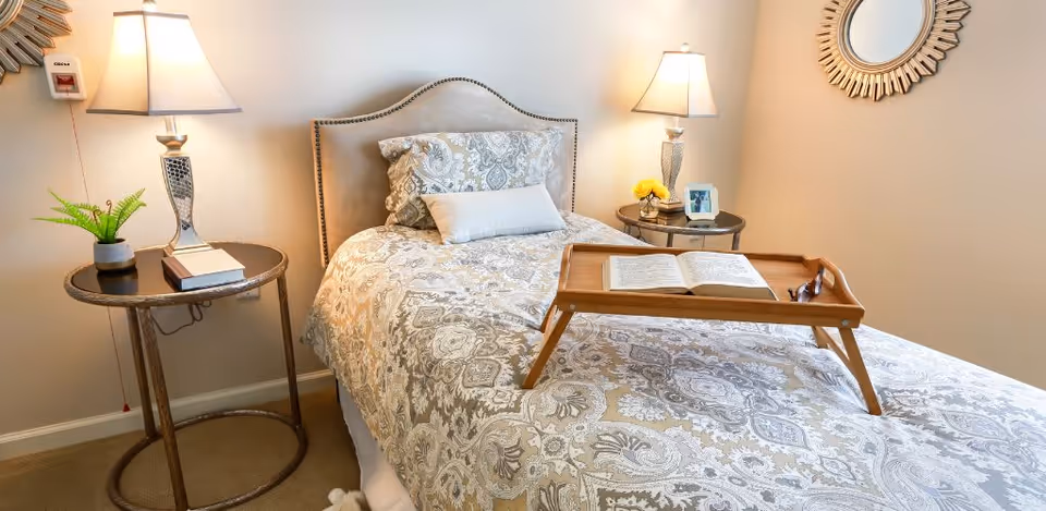 Neatly made single bed with patterned bedding, a wooden reading tray holding an open book, and bedside tables with lamps and decor.