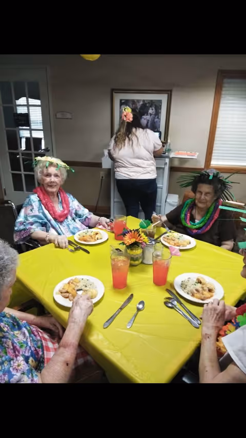 Four elderly women seated around a table covered with a yellow tablecloth, enjoying a meal with plates of food and glasses of pink beverage. They are wearing colorful leis and festive headbands. A woman stands in the background facing a computer on a small desk. The room has beige walls, a window with blinds, and a framed picture on the wall.