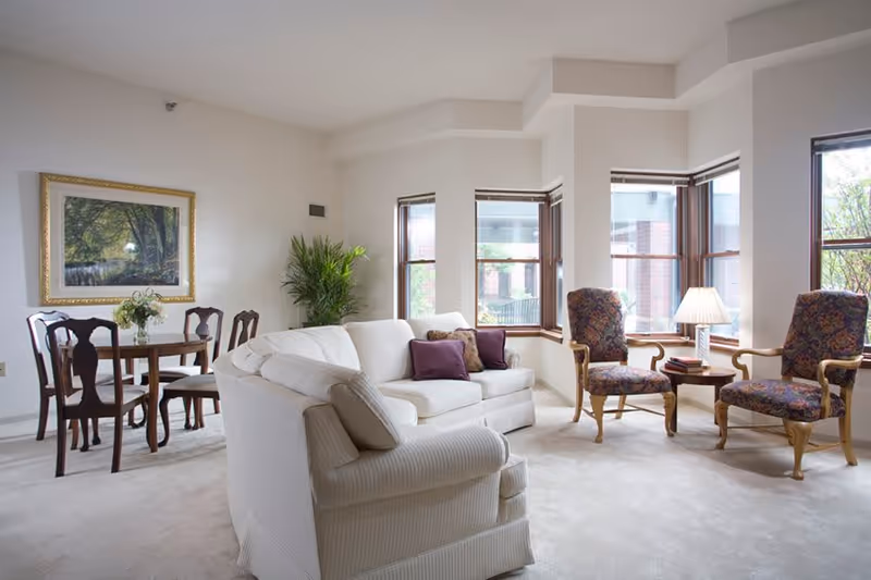 Bright living room with a white sofa, two upholstered chairs, a small dining table, bay windows, artwork, and a potted plant.