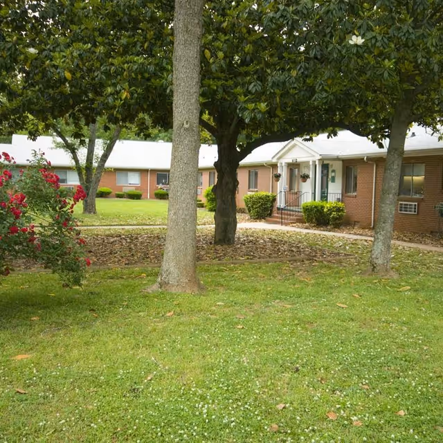 View of a single-story brick building with a white roof, surrounded by green grass, trees, and bushes. A concrete walkway leads to the entrance with a small porch and steps. There are flowering plants and well-maintained landscaping in the yard.