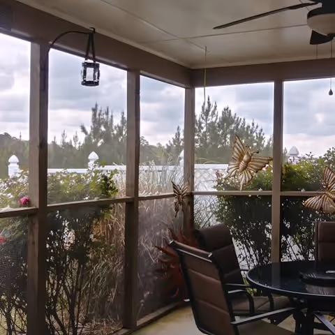 A screened-in porch area with a round glass table and several brown cushioned chairs. The porch is surrounded by mesh screens and wooden frames, with decorative butterfly ornaments attached to the screens. Outside the porch, there are green bushes and trees visible under a cloudy sky.