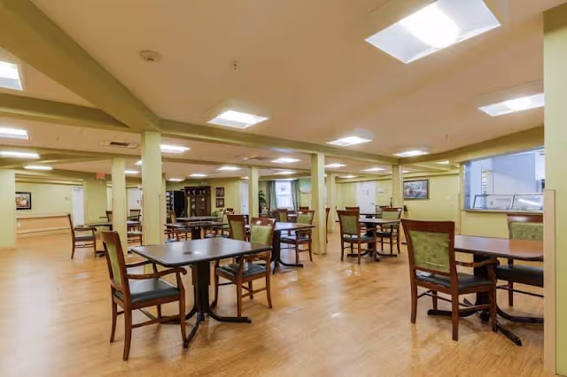 Large communal dining room with multiple square tables and wooden chairs on a hardwood floor under recessed ceiling lights.
