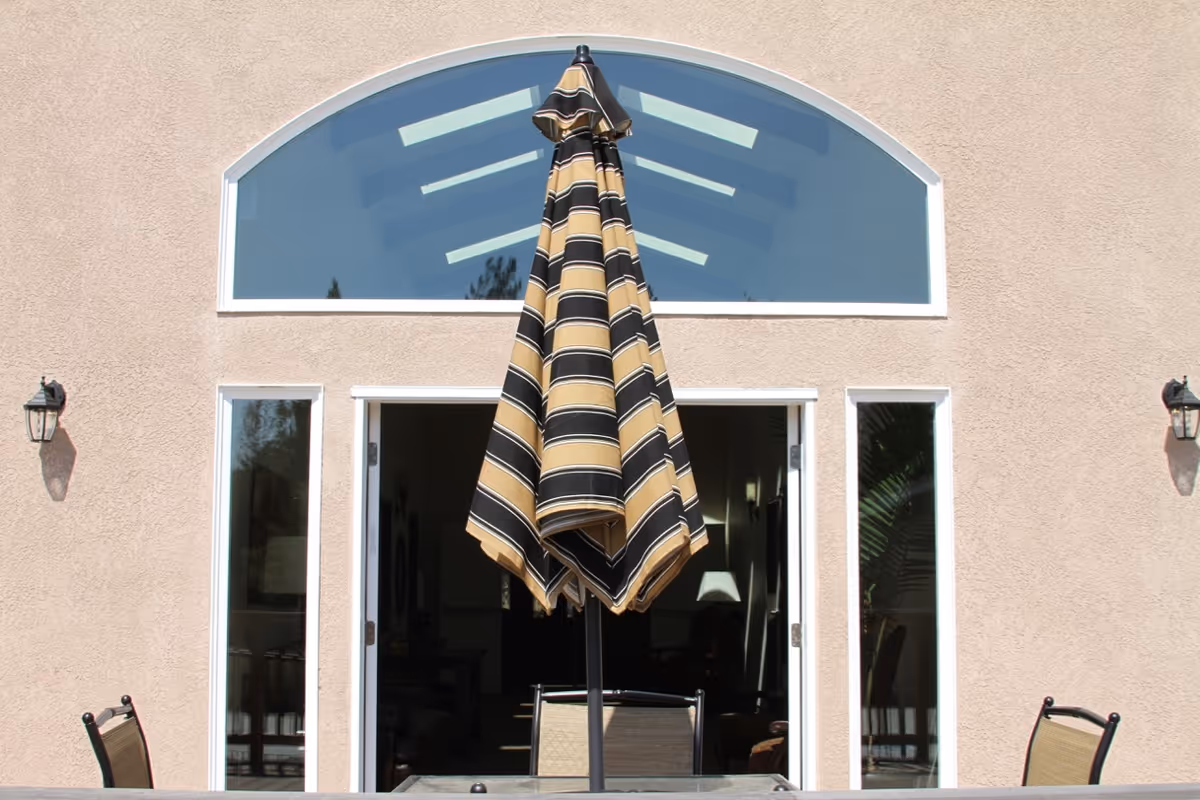 Outdoor patio area with a closed striped umbrella centered in front of large glass doors and windows on a beige building wall, with two outdoor wall lanterns on either side.