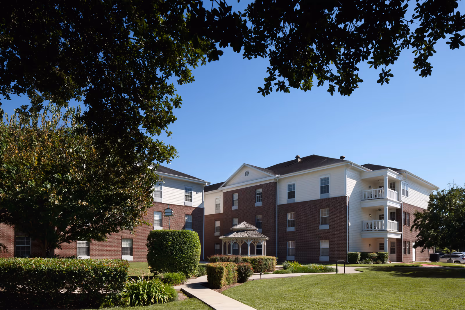 Three-story brick-and-siding senior living building with balconies, a small gazebo, manicured lawn and trees under a clear blue sky.